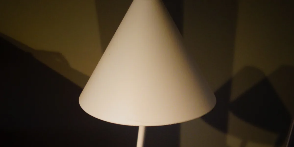 Weißer kegelförmiger Lampenschirm vor einer Wand, auf der Schatten in großen dunklen Flächen liegen