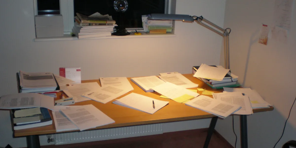 Schreibtisch am Abend voller bedruckter Blätter, Bücher und gelber Notizzettel, darüber eine silberne Tischlampe mit zweiteiligem Ausleger, dahinter ein dunkles Fenster mit gestapelten Zeitschriften