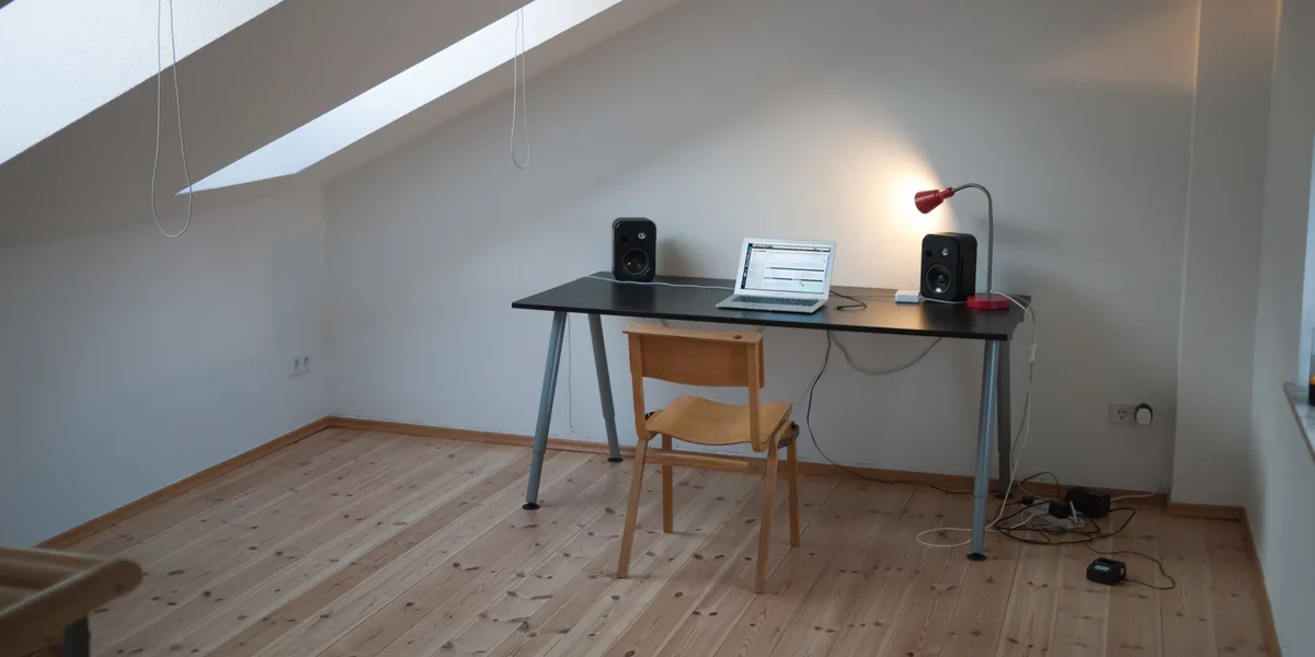 Schwarzer Tisch unter einer Dachschräge mit Laptop, zwei Lautsprechern und einer kleinen roten Tischlampe, deren Licht an die helle Wand fällt, davor ein Holzstuhl auf Dielenboden